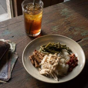 A plate of nasi campur with iced tea and rupiah notes on a wooden table in Bali